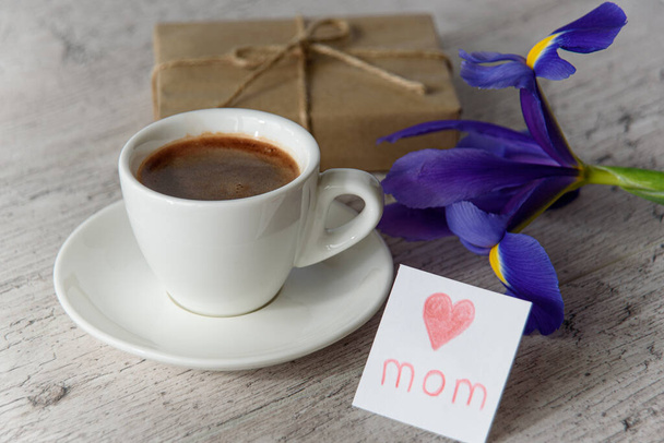 Fehér csésze kávé, kártya anyának a gyerektől, ajándékdoboz és íriszvirág a szürke fa asztalon. Boldog anyák napját! - Fotó, kép