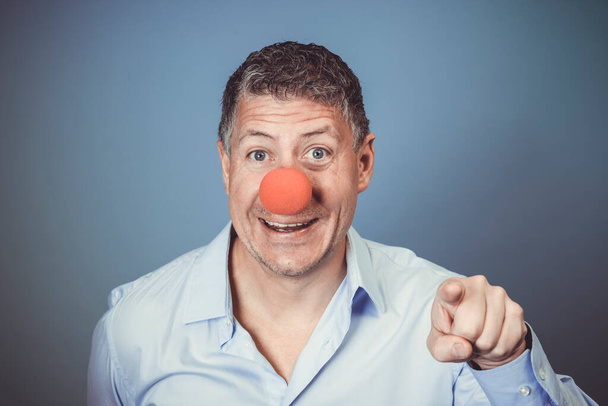 Middle aged man with blue shirt and red clown nose posing against blue background in the studio - Фото, зображення
