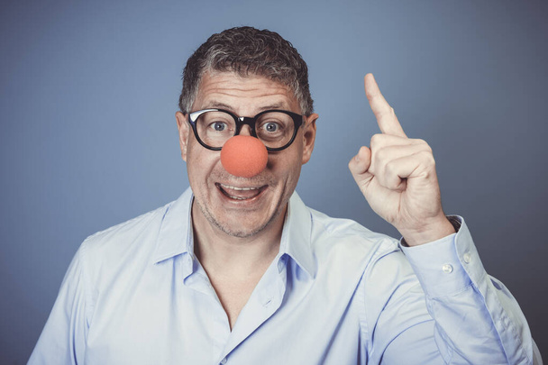 businessman with blue shirt and black glasses and red clown nose posing in front of blue background in the studio, pointing finger up - Φωτογραφία, εικόνα