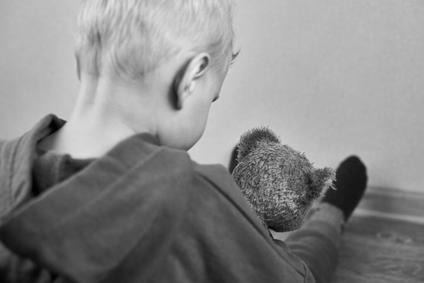 Ένα τρίχρονο καυκάσιο αγόρι κάθεται μόνο του στο πάτωμα και αγκαλιάζει αρκουδάκι, πίσω όψη, επιλεκτική εστίαση, το θέμα της παιδικής κακοποίησης, ασπρόμαυρη φωτογραφία. - Φωτογραφία, εικόνα