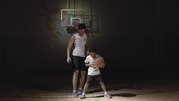 Handheld tracking shot of young male basketball player in sportswear teaching his little son how to dribble ball on dark indoor court - Footage, Video