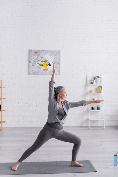 mujer de mediana edad en ropa deportiva de pie en pose guerrera mientras practica yoga - Foto, imagen
