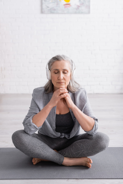 žena středního věku v bezdrátových sluchátkách meditující při sezení v lotosu pózovat na podložce jógy  - Fotografie, Obrázek