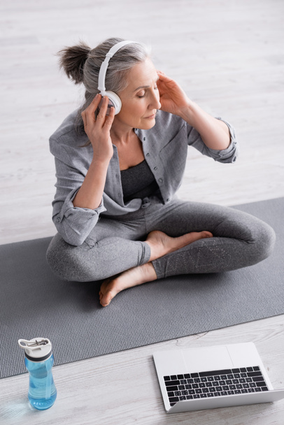 высокий угол зрения женщины средних лет в беспроводных наушниках медитирует, сидя в позе лотоса на коврик для йоги рядом с ноутбуком  - Фото, изображение