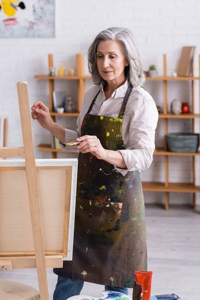 middle aged artist in apron holding paintbrush while painting at home - Photo, Image