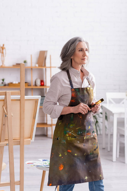 pleased middle aged artist in apron with spills holding paintbrush at home  - Foto, immagini