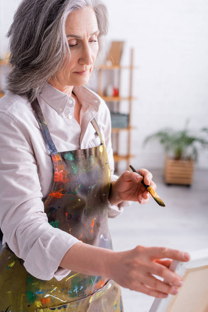 middle aged woman holding paintbrush near easel with canvas  - Foto, Bild