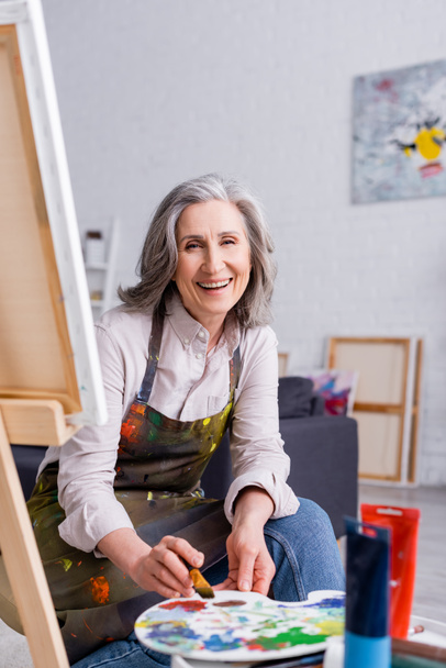 alegre artista de mediana edad sosteniendo pincel y paleta con pinturas de colores cerca del caballete con lienzo  - Foto, Imagen
