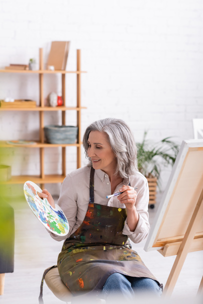 cheerful mature woman in apron holding paintbrush and palette near easel at home with blurred foreground  - Foto, Imagem