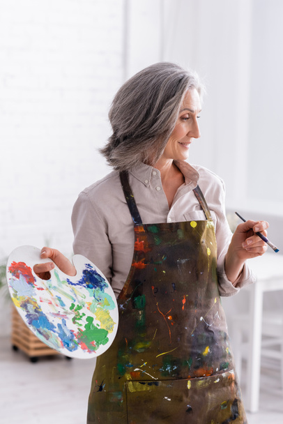 cheerful mature woman in apron holding paintbrush and palette  - Фото, изображение