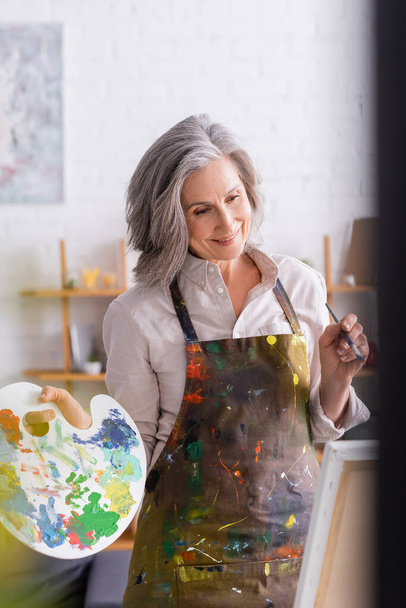 cheerful mature woman in apron holding paintbrush and palette while looking at canvas - Foto, immagini