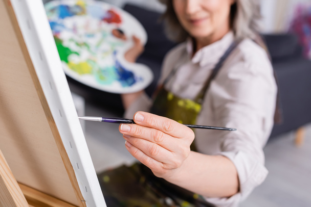 vista parcial de la mujer madura sosteniendo el pincel mientras pinta sobre lienzo con el primer plano borroso  - Foto, Imagen