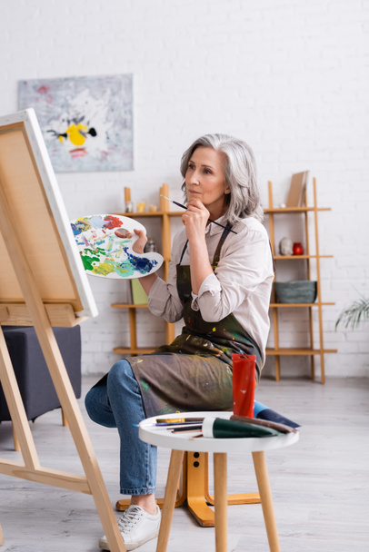 artista maduro pensativo sosteniendo pincel y paleta mientras está sentado cerca de la lona  - Foto, Imagen