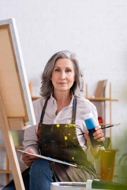 zralý umělec drží trubici s modrou barvou a paletou, zatímco sedí v blízkosti plátna  - Fotografie, Obrázek