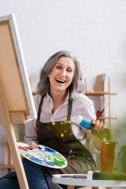 happy mature artist holding tube with blue paint and palette while sitting near canvas  - Photo, Image