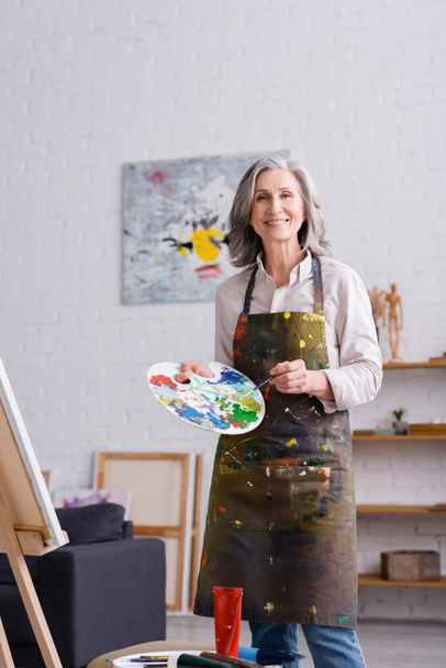 mujer de mediana edad positiva sosteniendo pincel y paleta con pinturas de colores cerca del caballete  - Foto, Imagen