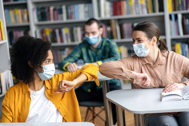 Two female student friends wearing protective face masks greeting each other bumping elbows while sitting at university library, healthcare concept, precautions, new normal - Foto, Bild