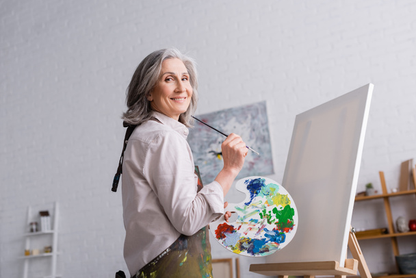 cheerful middle aged woman holding paintbrush and palette with colorful paints near blank canvas - Fotoğraf, Görsel