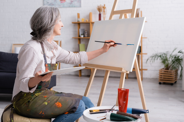 mujer alegre de mediana edad sosteniendo pincel y paleta mientras pinta sobre lienzo - Foto, Imagen