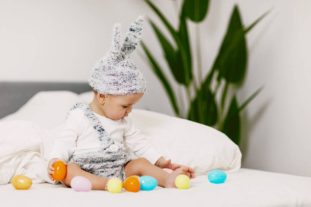 Dziecko siedzi na łóżku w stroju króliczka i bawi się kolorowymi pisankami, śmieje się i bawi - Zdjęcie, obraz