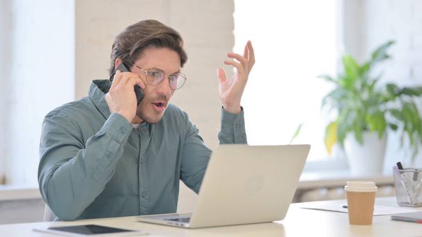 Dühös ember laptoppal beszél okostelefonon - Fotó, kép