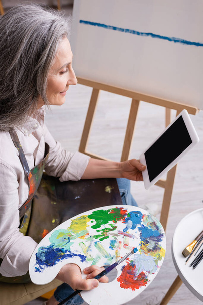 mature woman holding palette and digital tablet with blank screen near canvas - Фото, зображення