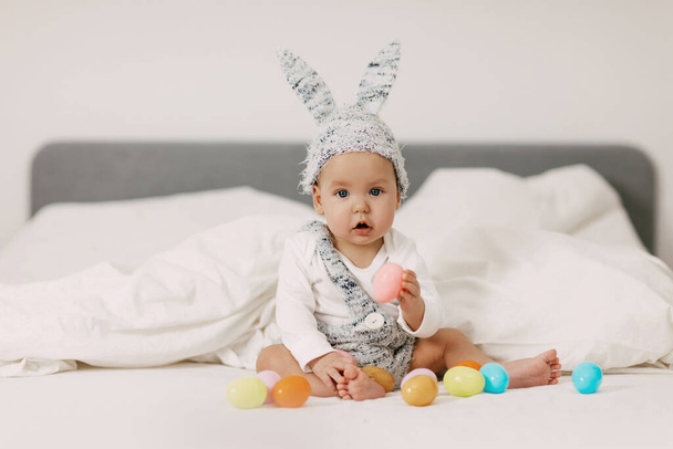 Дитина сидить на ліжку в кролячому костюмі і бавиться з барвистими пасхальними яйцями, сміється і розважається. - Фото, зображення