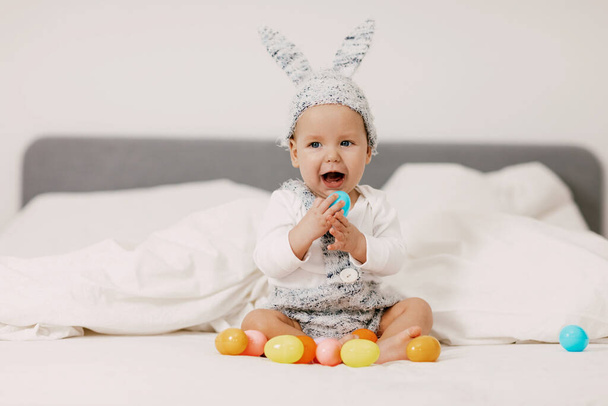 Baby zit op een bed in een konijnenkostuum en speelt met kleurrijke paaseieren, lacht en heeft plezier - Foto, afbeelding