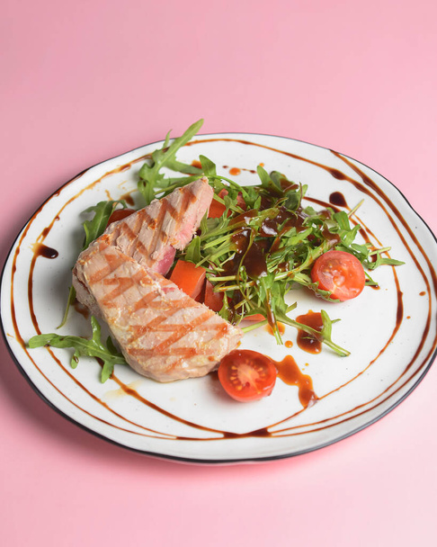 Close up still life of a tuna steak with fresh vegetables and arugula with seasoning over pastel pink background. Served on white plate with a fork. Delicious lunch or dinner, Italian cuisine. - Fotoğraf, Görsel