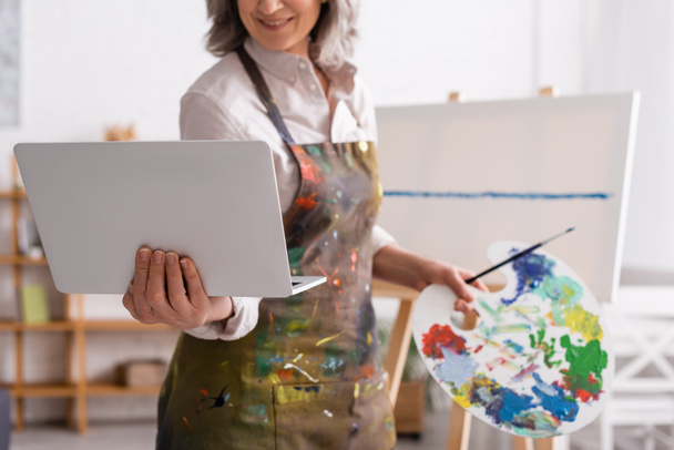 cropped view of mature woman holding palette and laptop while watching tutorial near canvas - Foto, Imagen