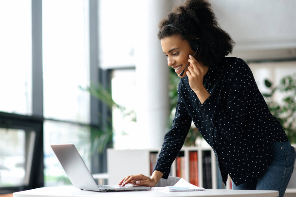 Busy successful African American young business woman, manager or consultant, in a headset, stands near a desktop in an office, uses a laptop, typing a message, or communicating with a client online - 写真・画像