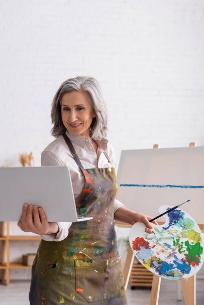 mosolygós érett nő gazdaság paletta és laptop, miközben néz tutorial közel vászon - Fotó, kép