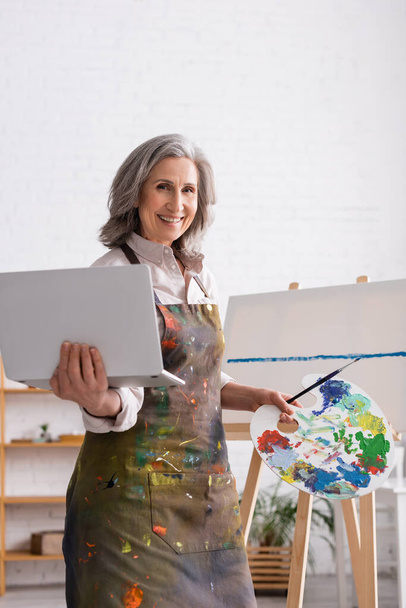 joyful mature woman holding palette and laptop near canvas - Photo, Image
