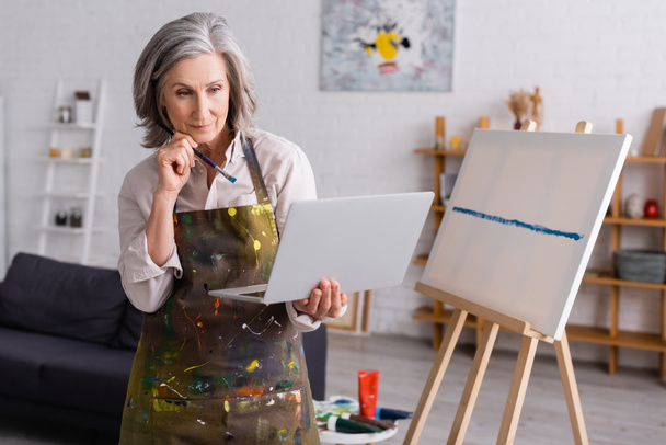 mature woman holding paintbrush and laptop while watching tutorial near canvas - Photo, Image