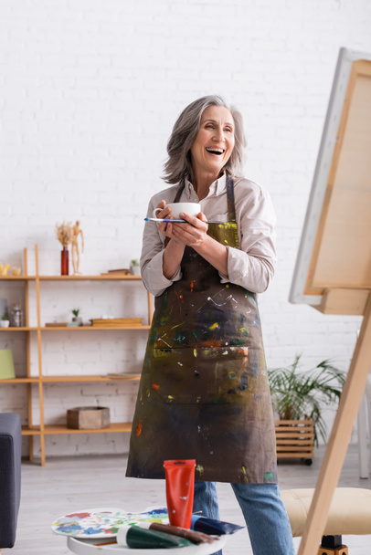 excited middle aged artist holding cup of coffee and paintbrush near canvas  - Photo, Image