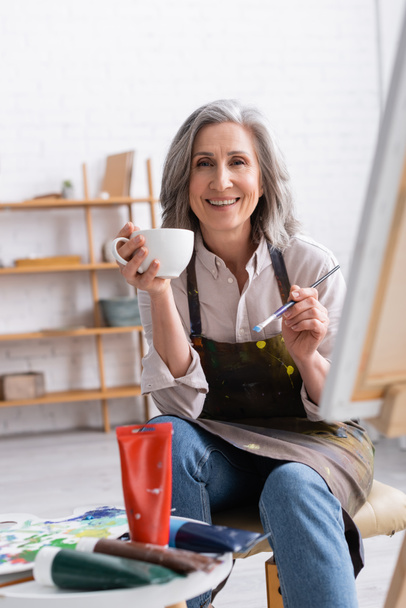 happy middle aged artist holding cup of coffee and paintbrush near canvas  - Foto, imagen