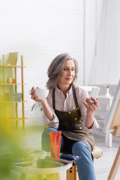 middle aged artist holding paintbrush, cup of coffee and using smartphone near canvas and blurred foreground - Photo, Image
