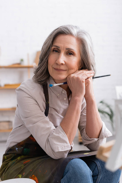 pleased middle aged woman with grey hair holding paintbrush  - Fotografie, Obrázek