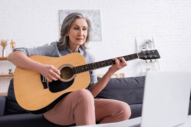 mujer de mediana edad con pelo gris aprendiendo a tocar la guitarra acústica cerca de la computadora portátil en primer plano borrosa  - Foto, imagen