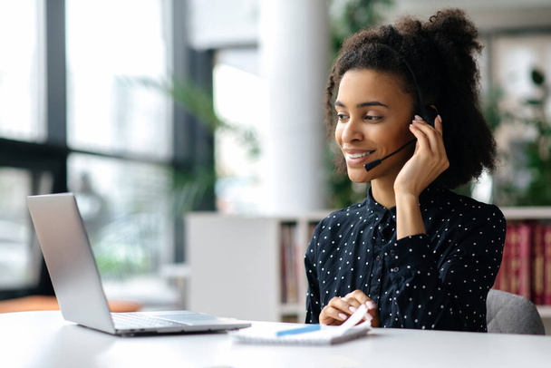 African American friendly young woman with headset, call center worker, consultant, business person, looks at the laptop screen, talking with colleagues or clients by video call, conducts consultation - Photo, image