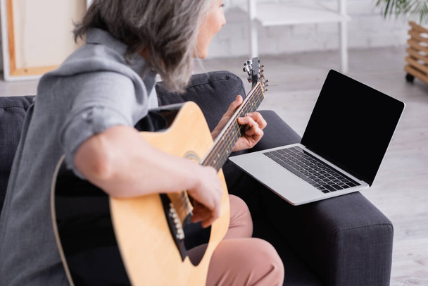 niewyraźne w średnim wieku kobieta uczy się grać na gitarze akustycznej w pobliżu laptopa z pustym ekranem na kanapie - Zdjęcie, obraz