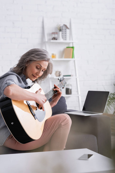 donna matura imparare a suonare la chitarra acustica vicino al computer portatile con schermo bianco sul divano  - Foto, immagini