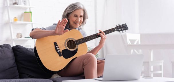 keski-ikäinen nainen harmaat hiukset tilalla akustinen kitara kun heiluttaa käsi videopuhelun aikana kannettavan tietokoneen, banneri - Valokuva, kuva