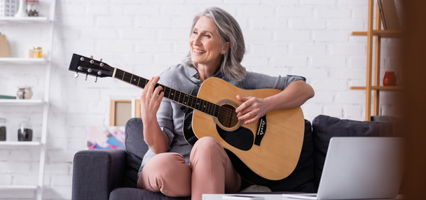 tyytyväinen kypsä nainen harmaat hiukset oppia soittamaan akustista kitaraa läppärin lähellä, banneri - Valokuva, kuva