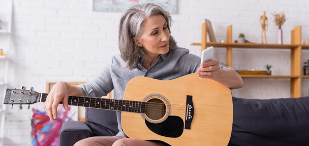 kypsä nainen, jolla on harmaat hiukset, katselee älypuhelinta oppien soittamaan akustista kitaraa, banneria - Valokuva, kuva