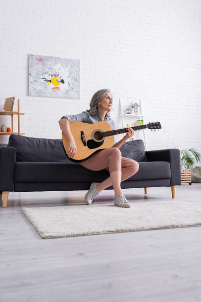 dreamy mature woman with grey hair sitting on couch with acoustic guitar  - Photo, Image