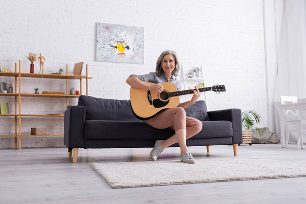 mulher madura alegre com cabelos grisalhos sentado no sofá e tocando guitarra acústica na sala de estar - Foto, Imagem