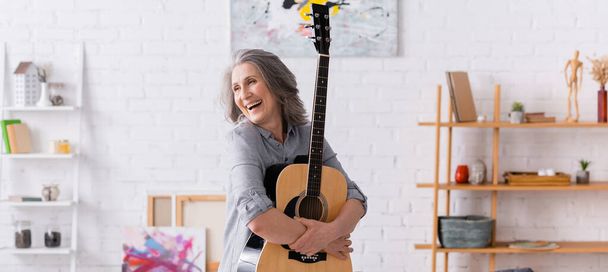 iloinen kypsä nainen harmaat hiukset seisoo akustinen kitara olohuoneessa, banneri - Valokuva, kuva