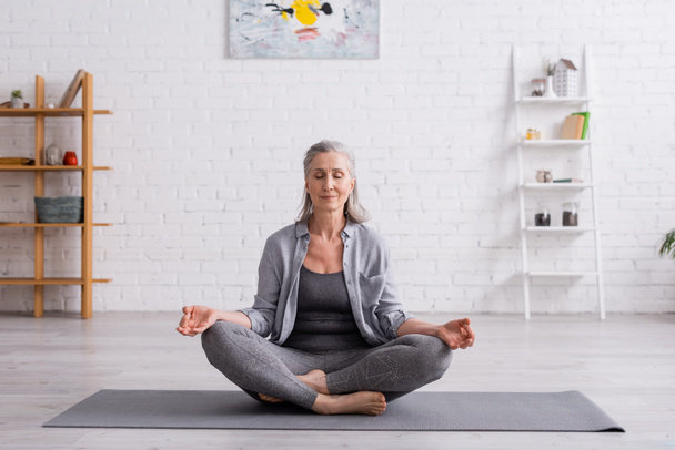mujer madura con pelo gris sentada en pose de loto en esterilla de yoga - Foto, Imagen