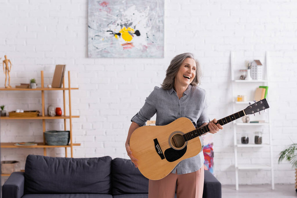 happy mature woman with grey hair standing with acoustic guitar and laughing in living room  - Foto, immagini